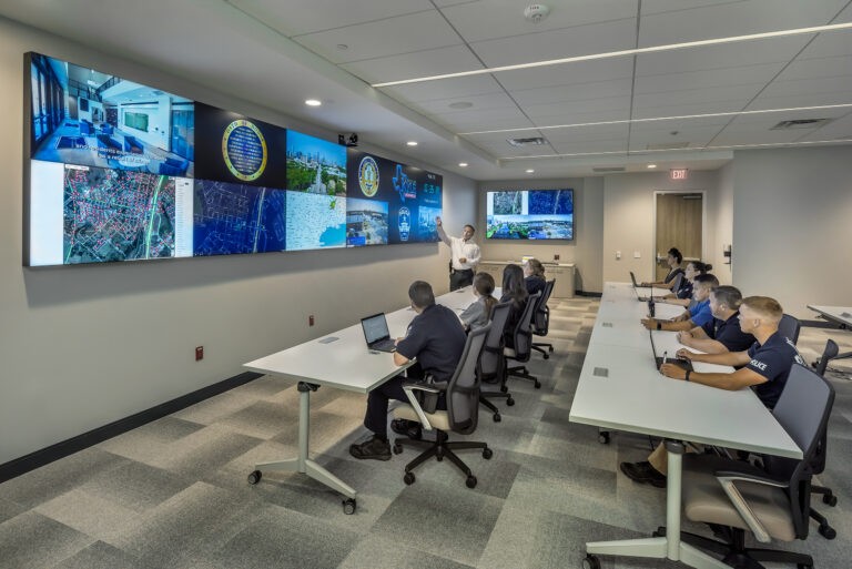 a group of police officers sit at tables in front of a large screen watching a presentation