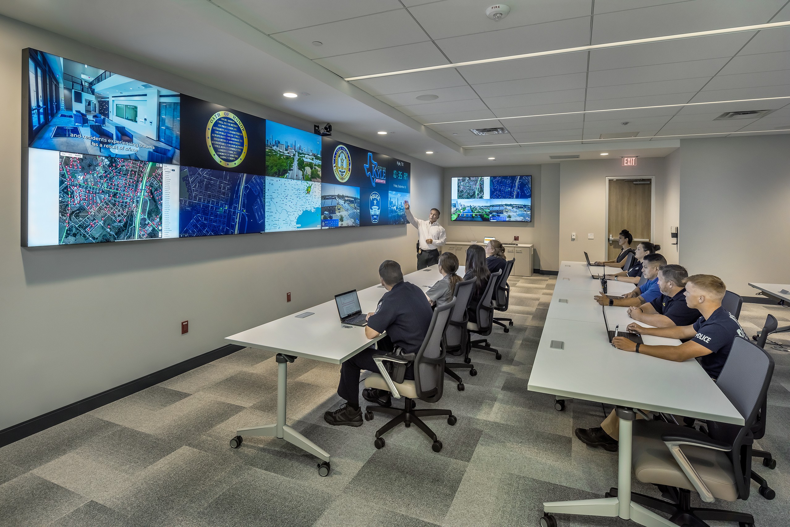 a group of police officers sit at tables in front of a large screen watching a presentation